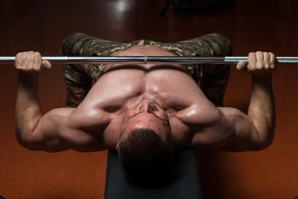 Homem musculoso sem camisa, com calça militar fazendo supino no banco da academia